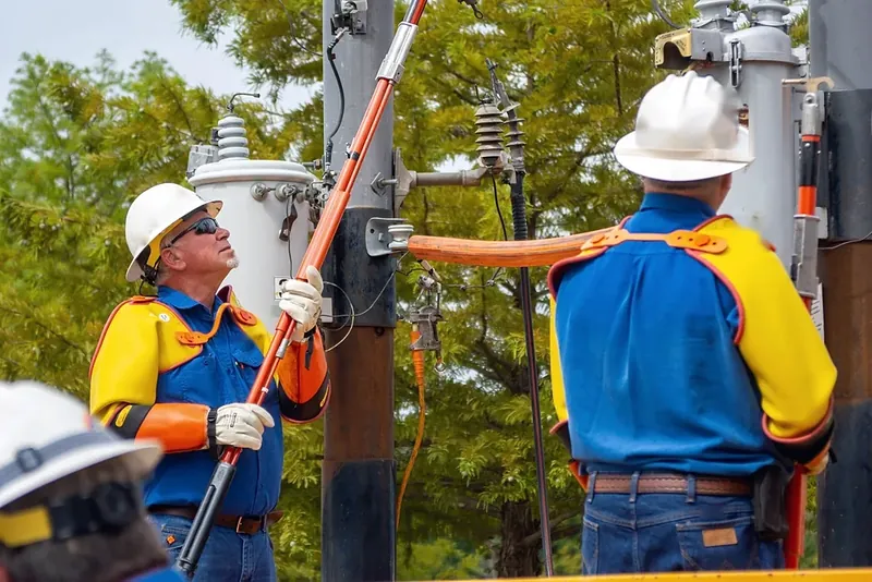 Linieros con casco y guantes dieléctricos maniobran una pértiga aislada junto a un transformador.